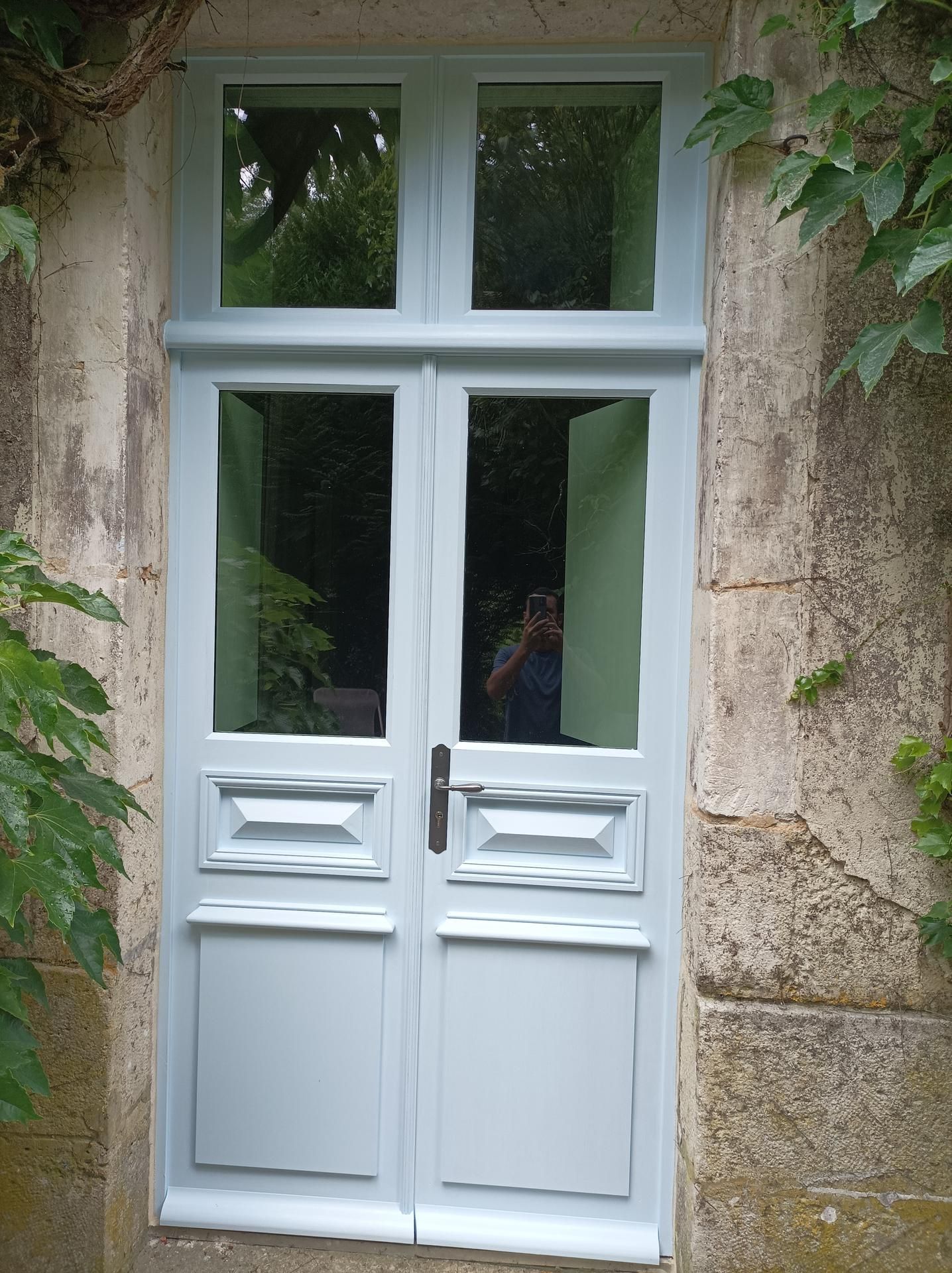 Double porte bleu pâle à panneaux de verre dans un encadrement en pierre, entourée de feuillage vert.