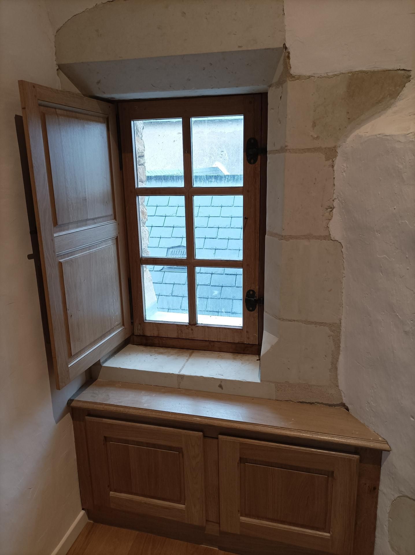 Fenêtre en bois aux volets ouverts, encastrée dans un mur de pierre. Un meuble se trouve sous la fenêtre.