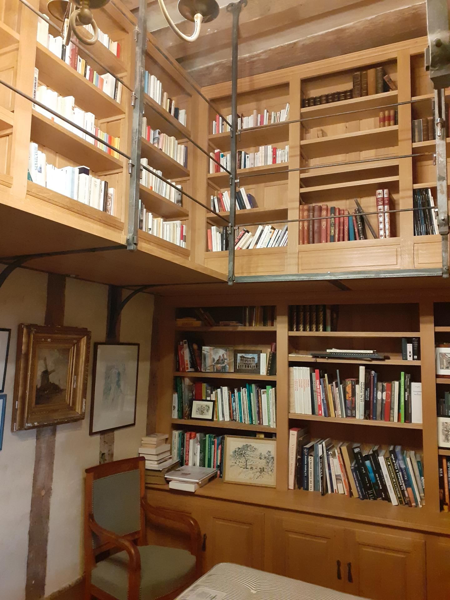 Des étagères en bois remplies de livres s'étendent sur deux niveaux, avec une chaise et des œuvres d'art au premier plan.