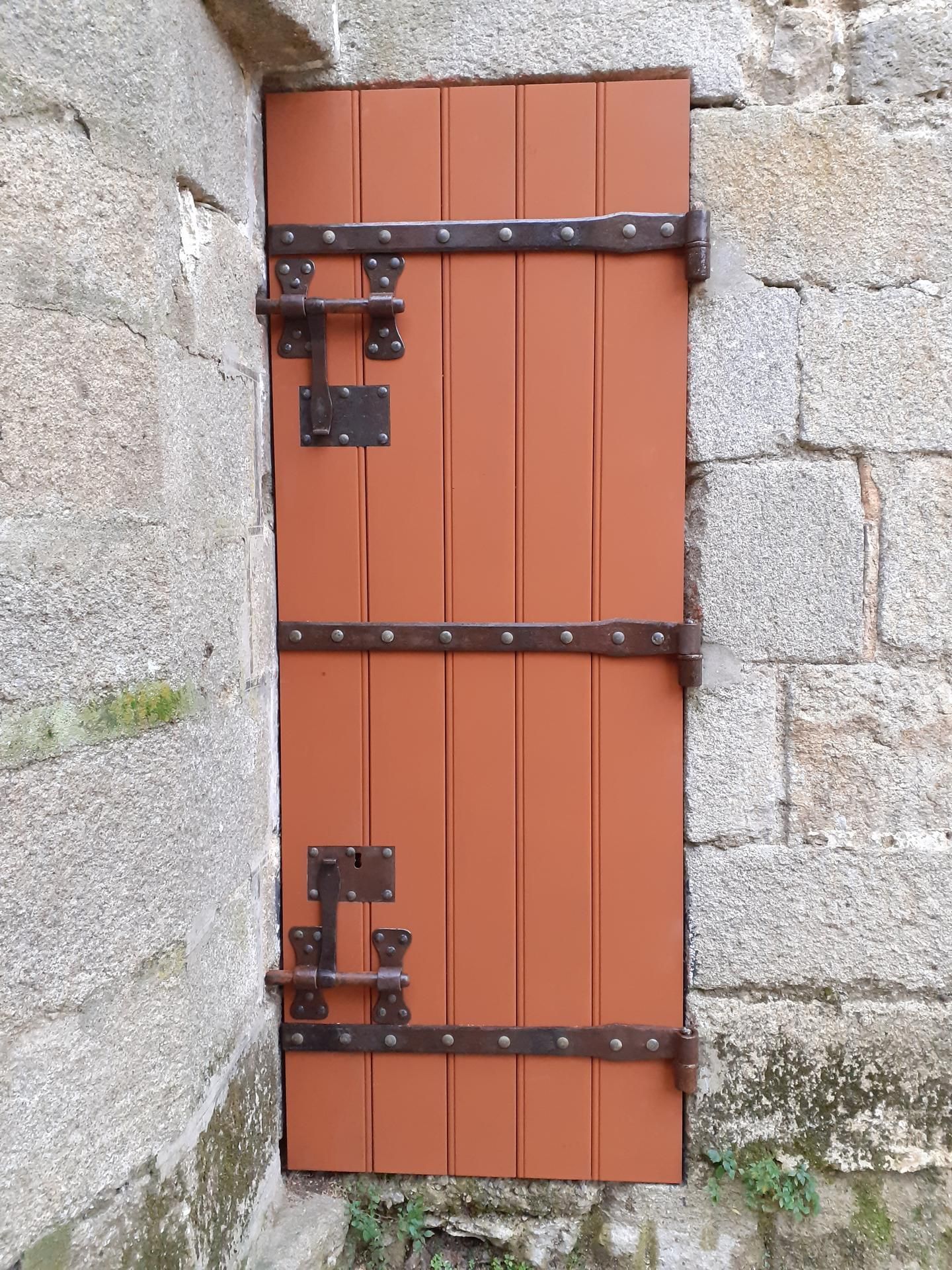 Porte en bois brun avec quincaillerie en métal foncé, encastrée dans un mur en pierre.