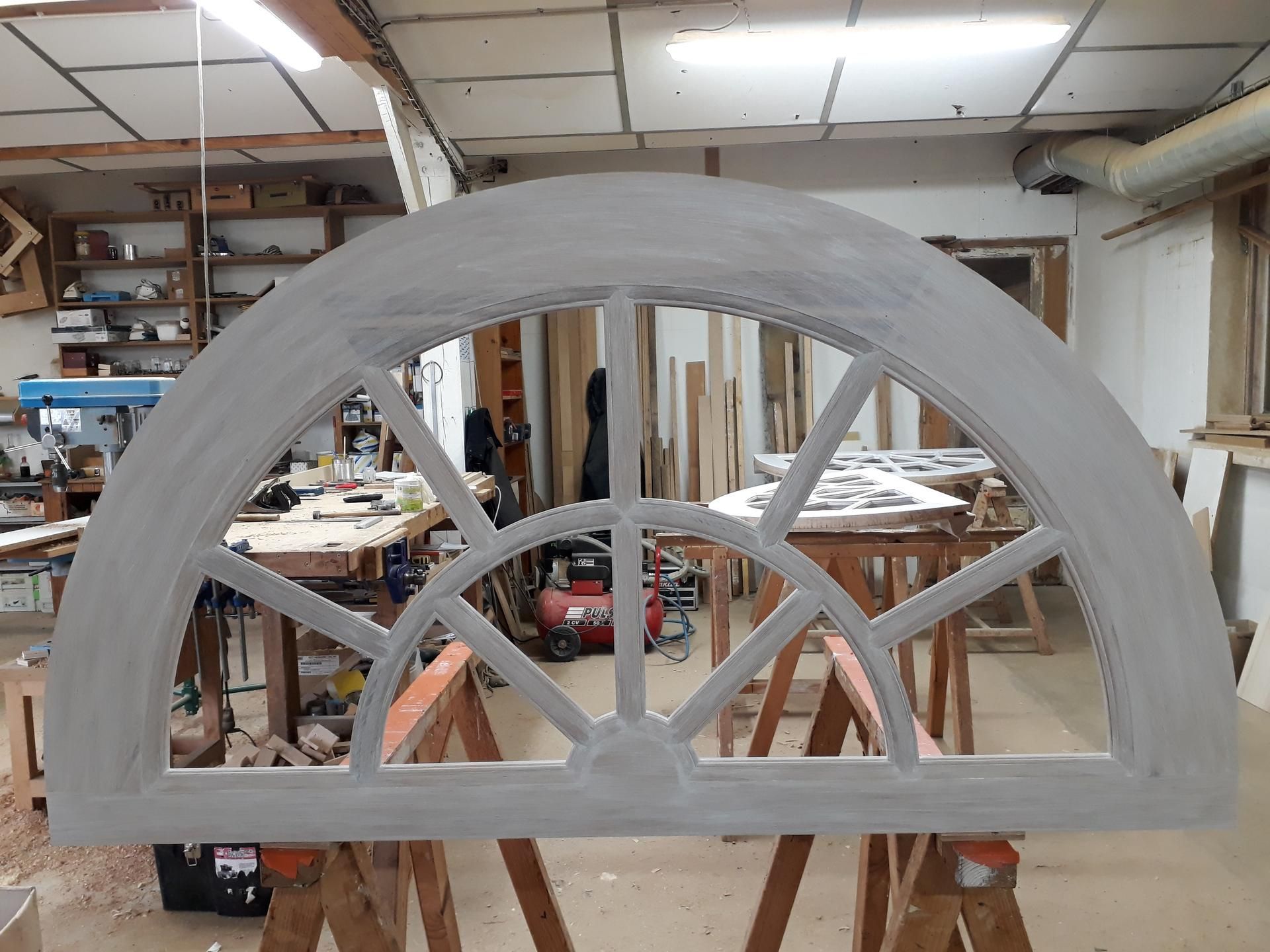 Un cadre de fenêtre en demi-cercle à plusieurs carreaux, peint en gris clair, dans un atelier.