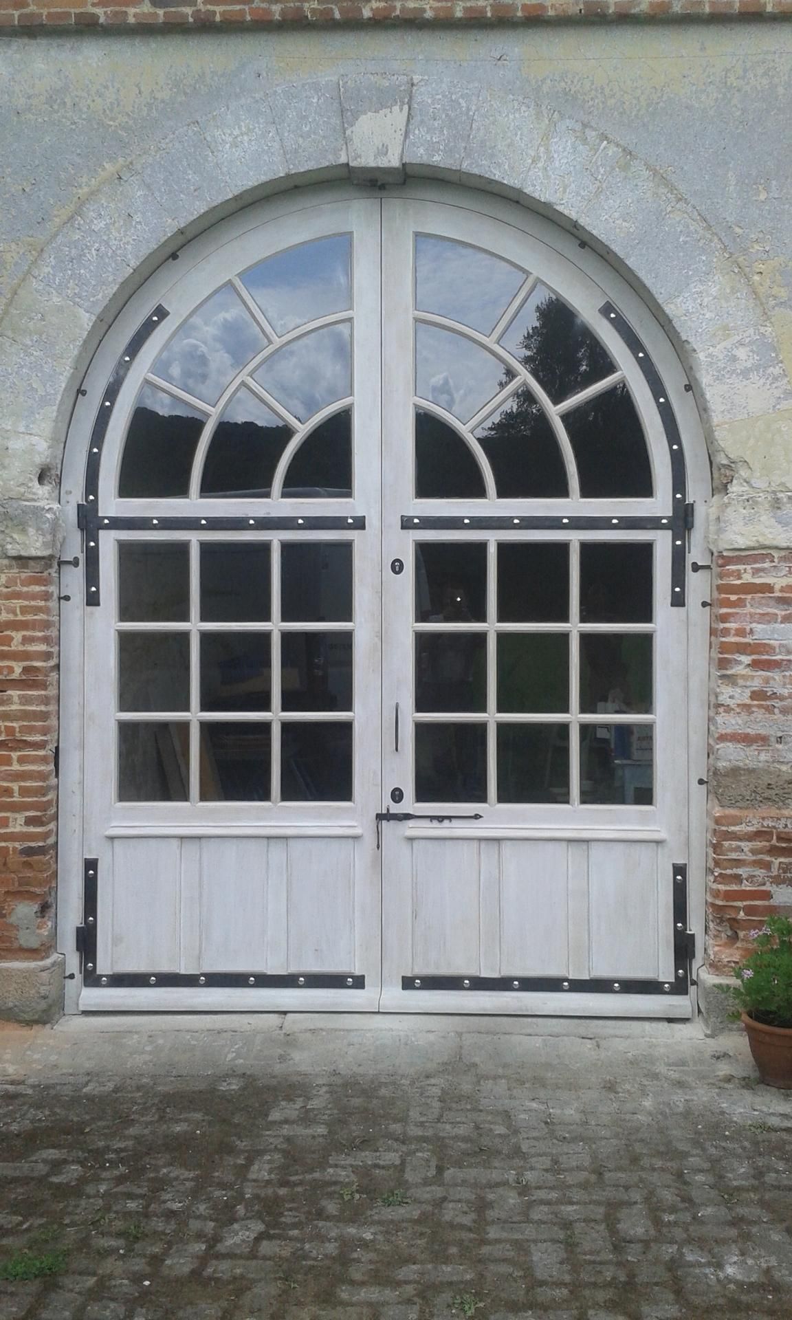 Double porte cintrée blanche avec panneaux de verre et charnières noires, intégrée à une façade en briques et en pierres.