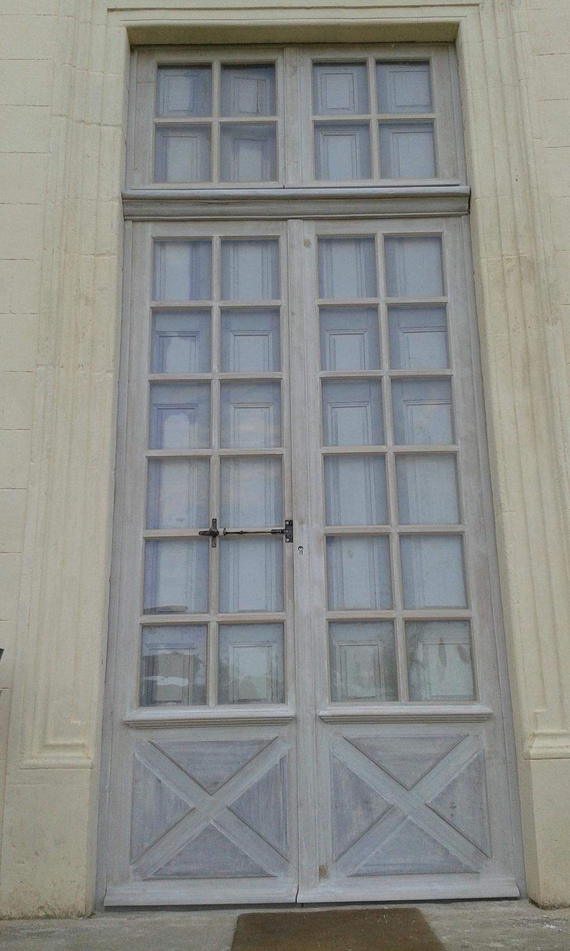 Vue extérieure de portes-fenêtres à plusieurs carreaux, patinées par le temps et munies d'impostes, sur une façade d'immeuble de couleur claire.