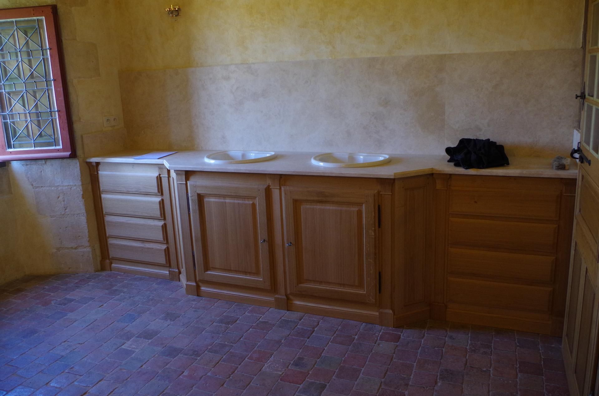 Salle de bains avec meubles en bois, lavabos et plan de travail en pierre. Sol en briques rouges, murs beiges.