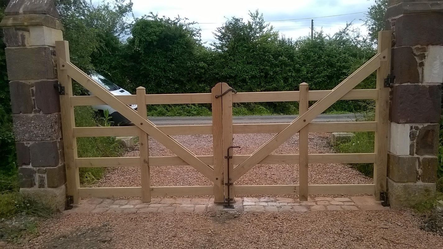 Portail de ferme en bois, brun clair, placé entre des colonnes de pierre, allée de gravier, fond de feuillage vert.