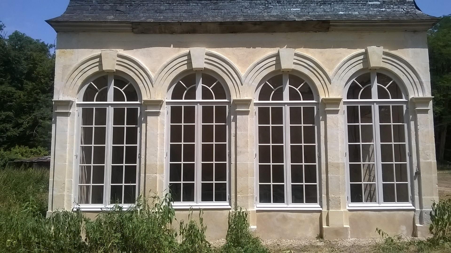 Bâtiment en pierre avec fenêtres cintrées, encadrements blancs, toit gris, entouré de verdure.