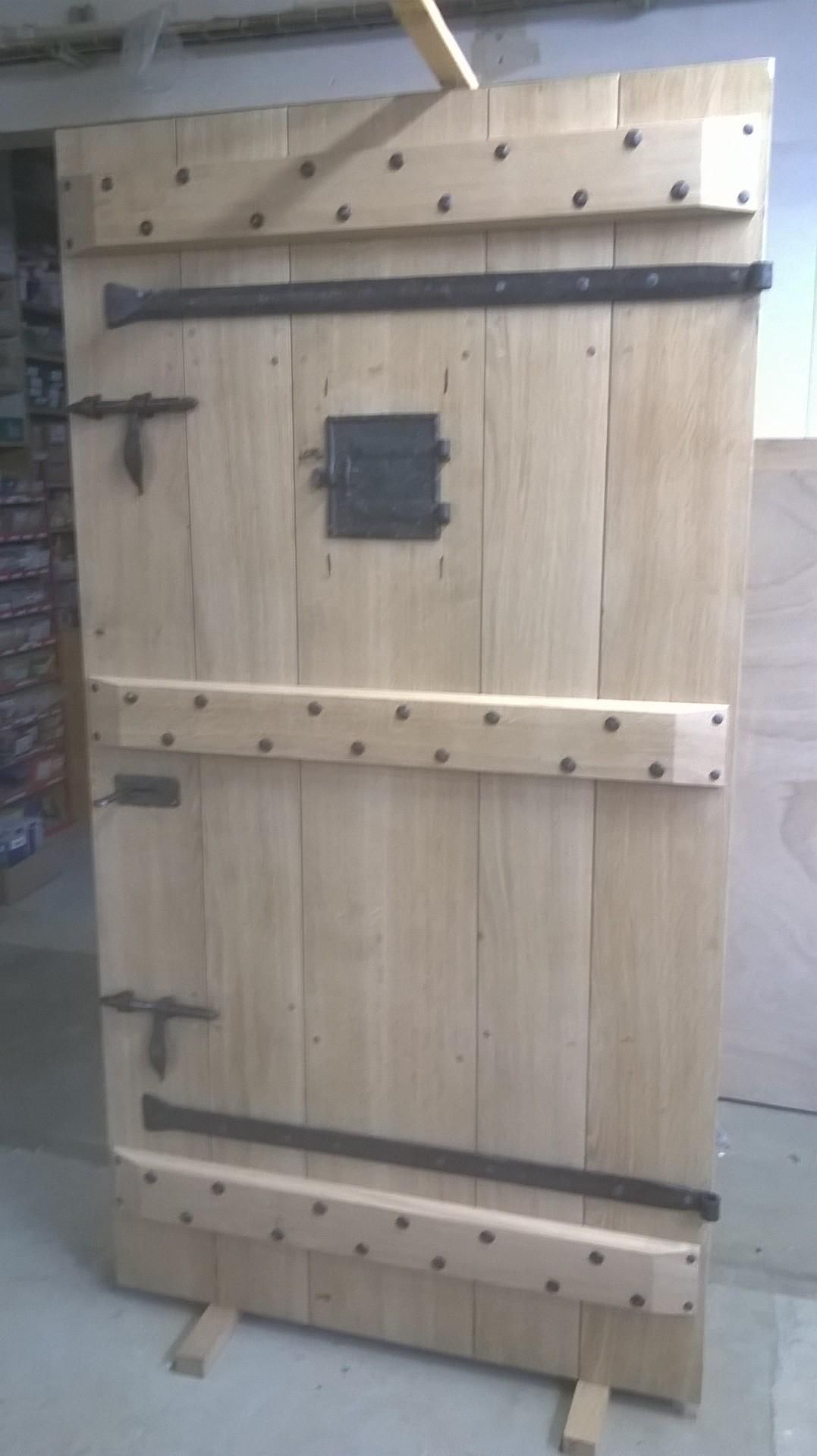 Porte en bois de style médiéval avec charnières et loquets en métal, et une petite ouverture carrée.