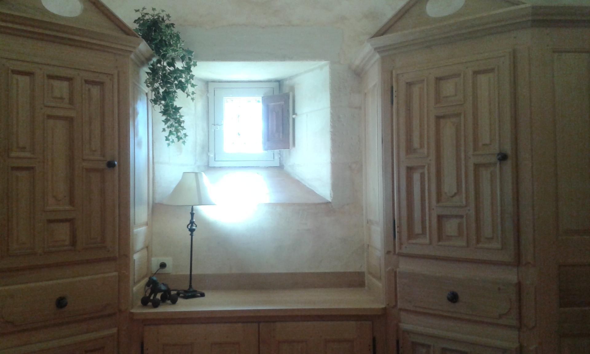 Des placards en bois intégrés encadrent une petite fenêtre où se trouvent une table, une lampe et des plantes décoratives.