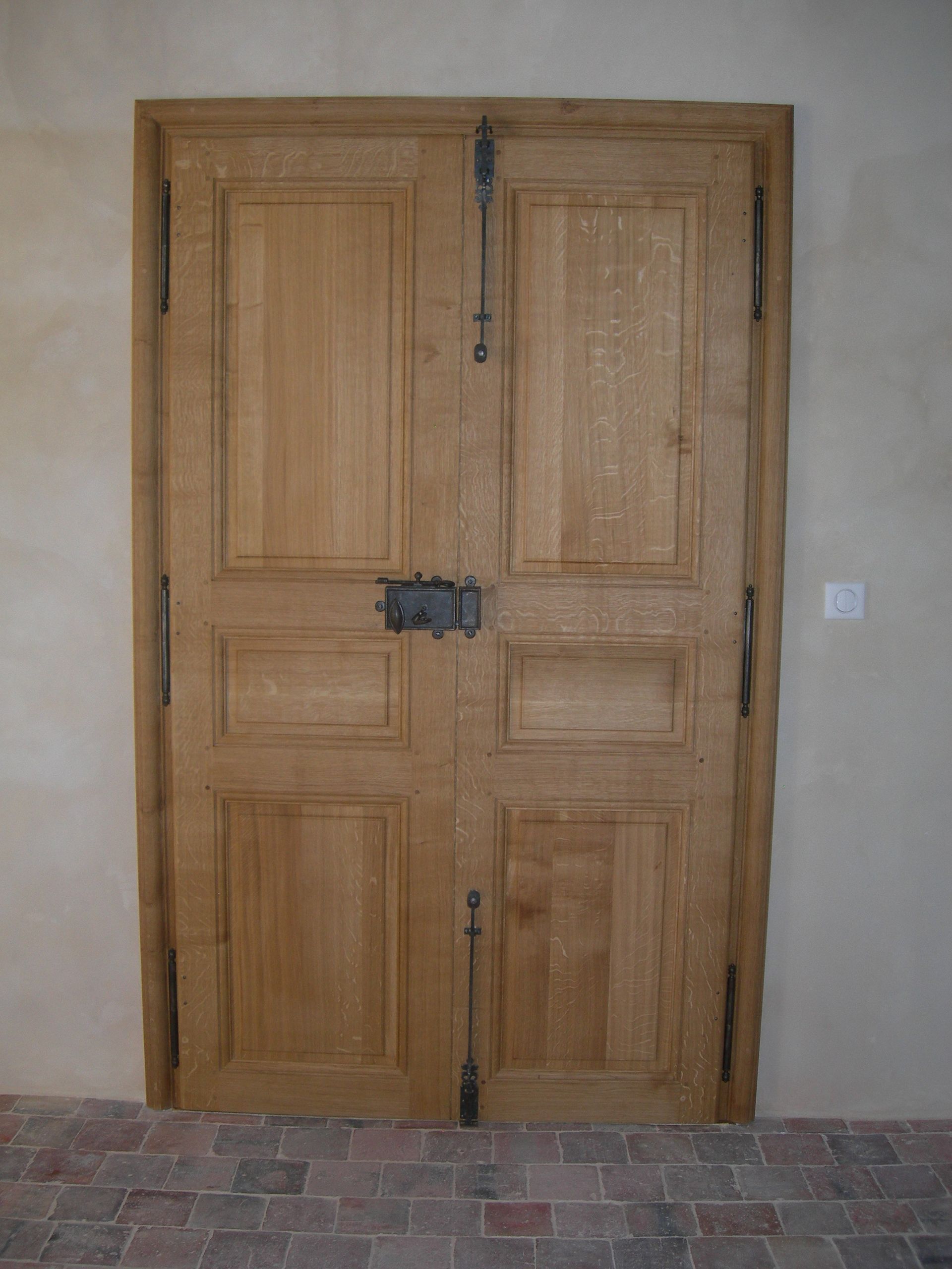 Double porte en bois avec quincaillerie foncée, encastrée dans un mur beige clair, sur un sol en briques.
