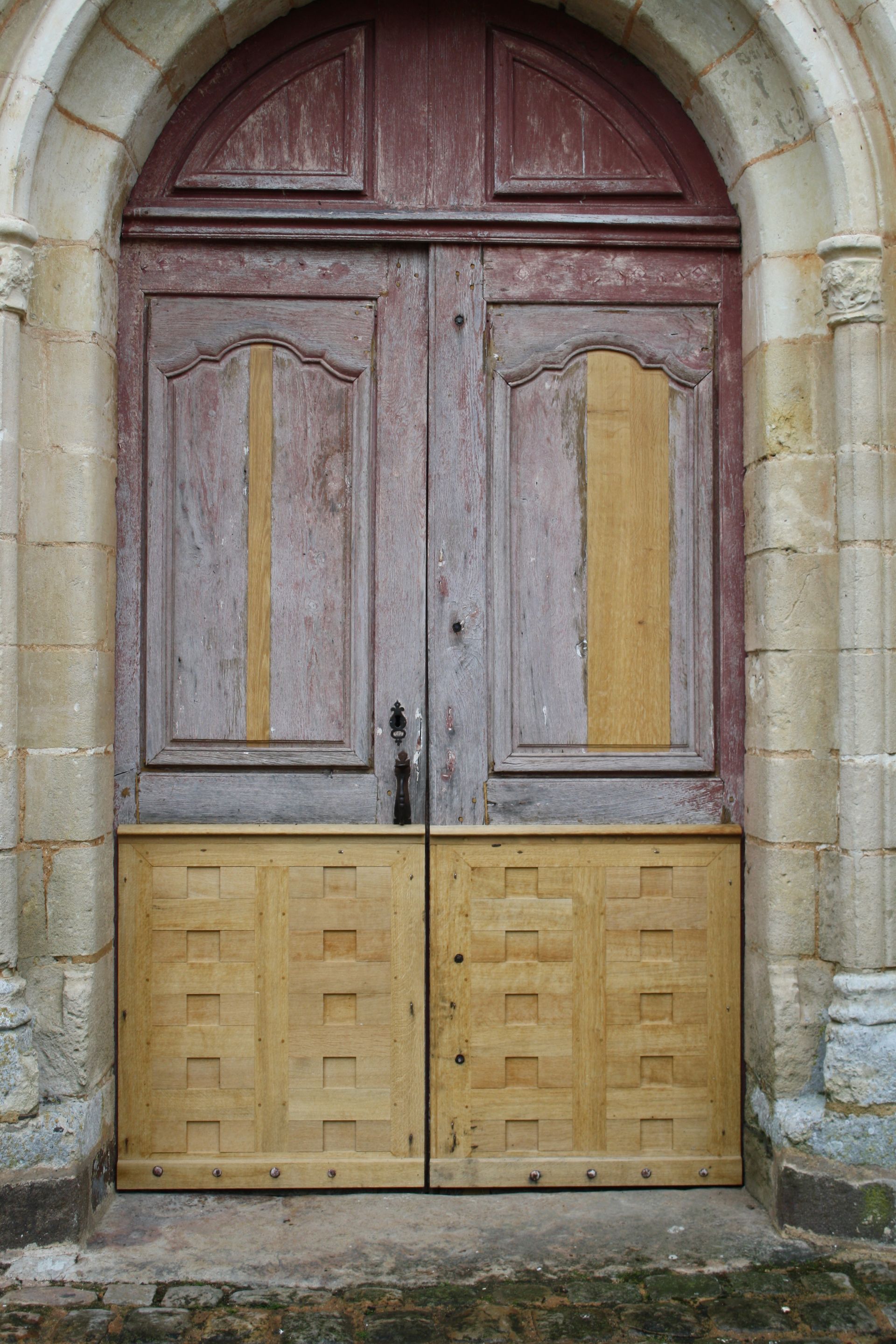 Vieilles portes d'église en bois patiné, peintes en beige et rouge, sous un cadre en pierre arqué.