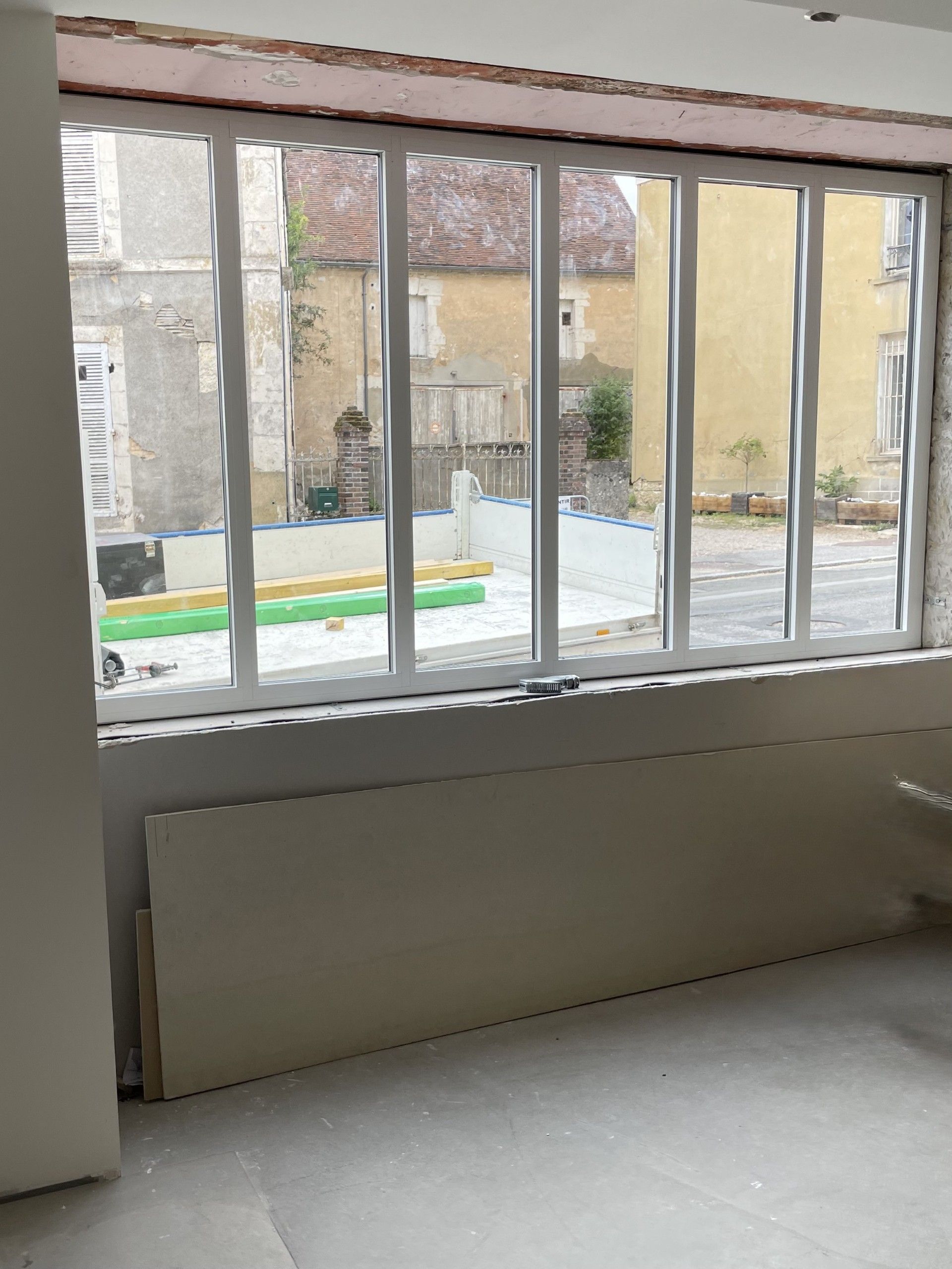 Vue intérieure d'une pièce avec une grande fenêtre donnant sur une cour intérieure bordée de vieux bâtiments. Boiseries blanches, murs clairs.