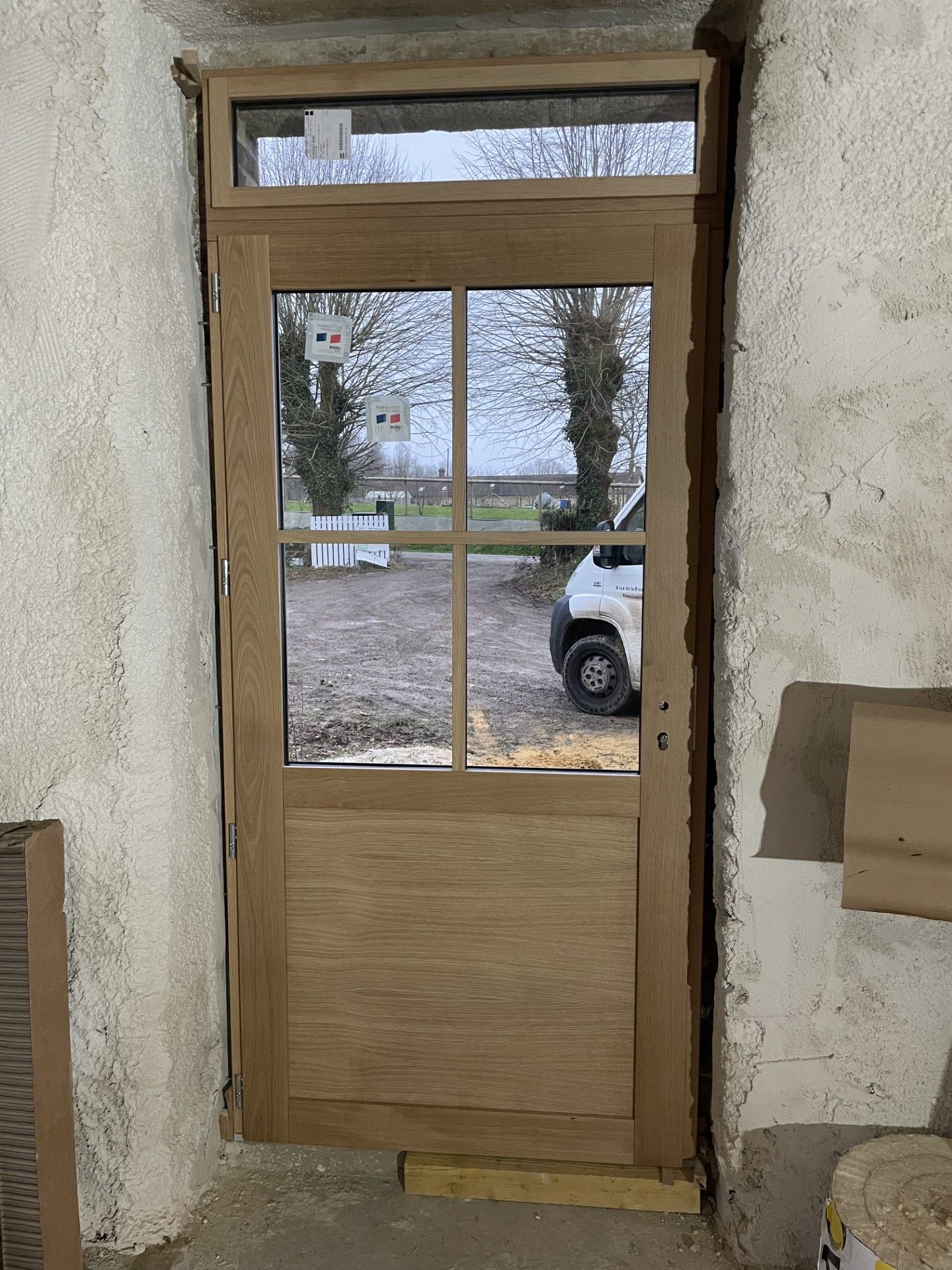 Porte en bois avec vitres et panneau de verre au-dessus, encastrée dans une entrée rustique et brute.