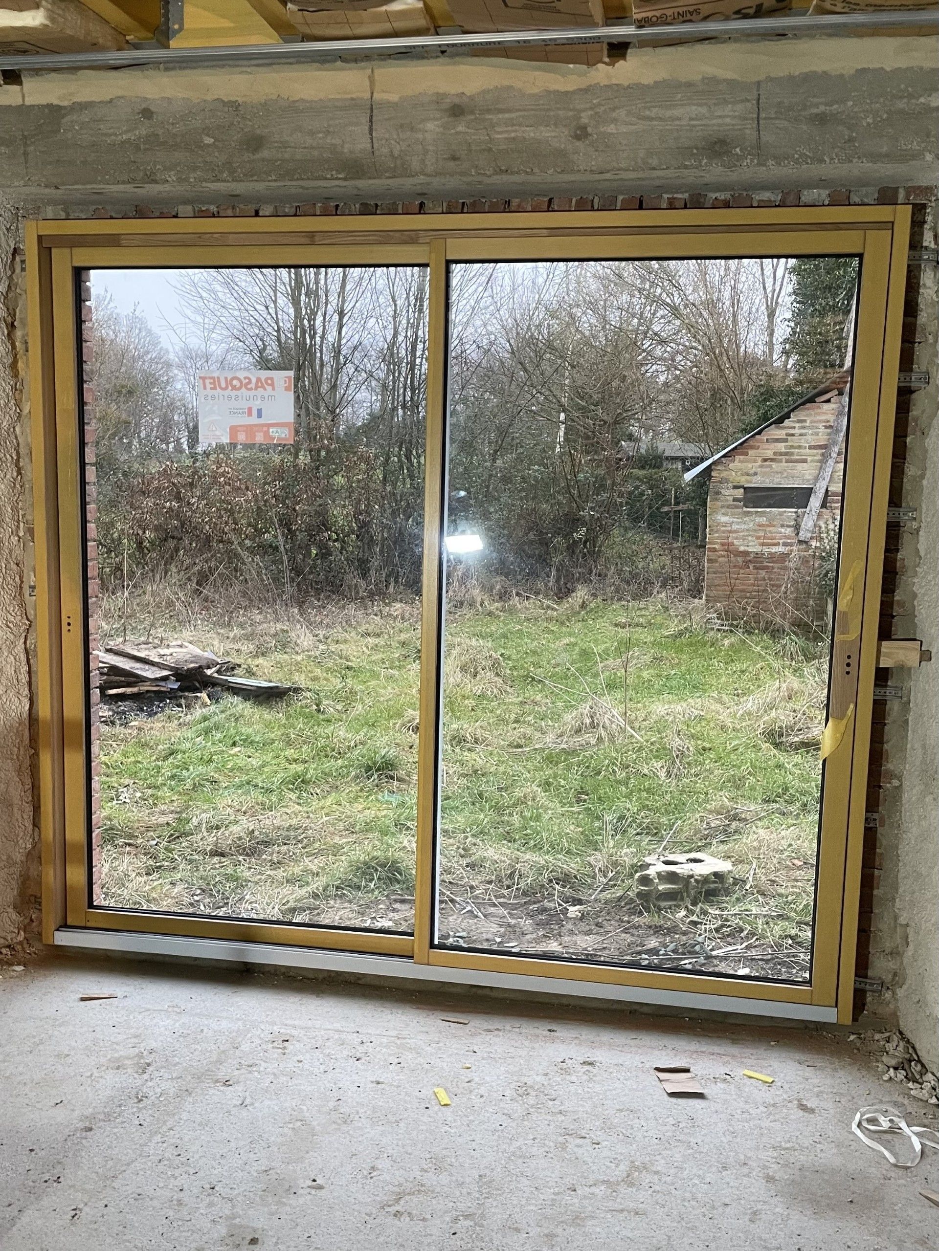 Porte coulissante en verre, encadrée de bois, donnant sur une cour arrière envahie par les herbes hautes, avec des arbres et un petit abri de jardin ; sol intérieur en béton.