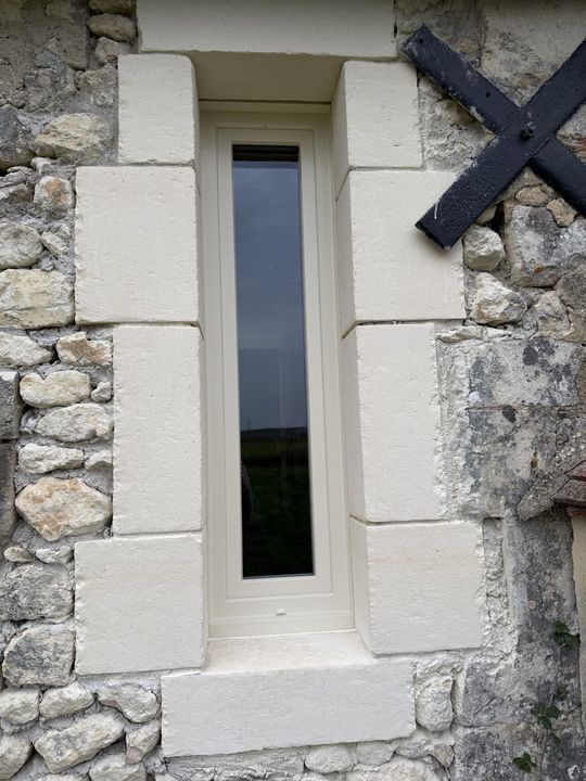 Étroite fenêtre dans un mur de pierre, entourée de blocs clairs. Entretoise noire visible.