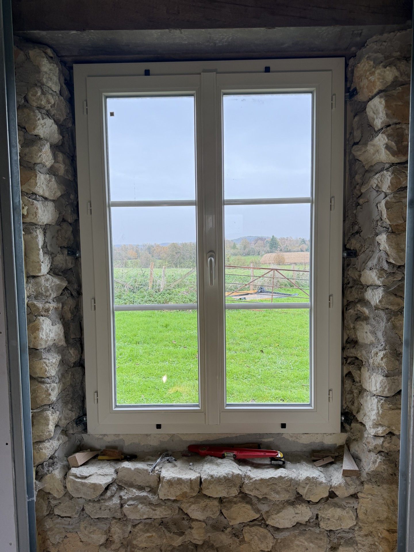 Fenêtre à cadre blanc dans un mur de pierre, donnant sur un champ verdoyant sous un ciel couvert.