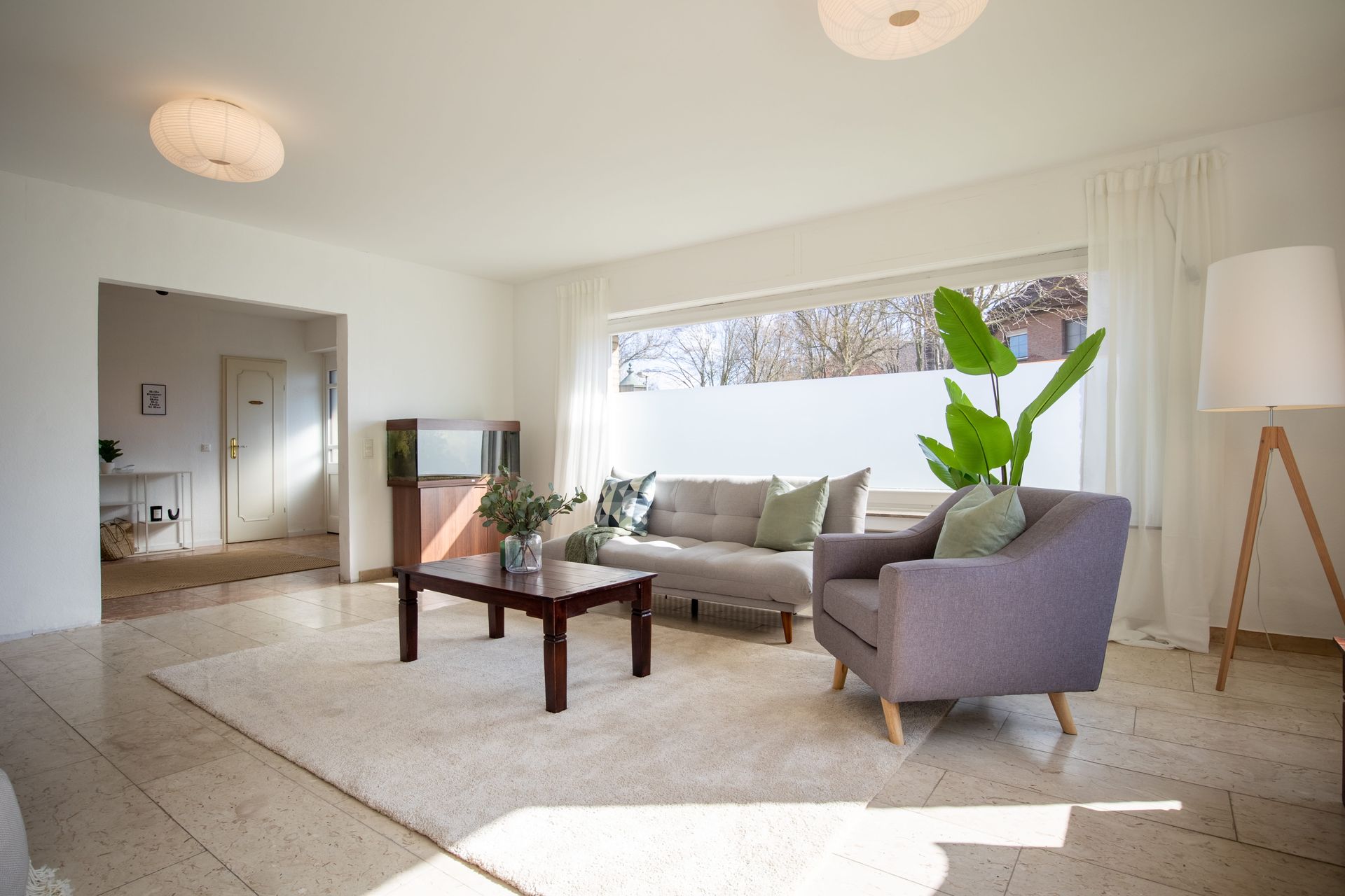 Ein helles Wohnzimmer mit beigefarbenem Teppich, grauem Sofa, lila Sessel, einem Couchtisch aus Holz.