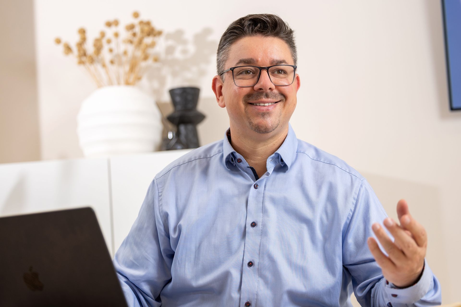 Eine Person mit Brille in einem hellblauen Hemd mit Knöpfen lächelt und gestikuliert während eines Gesprächs an einem Schreibtisch.