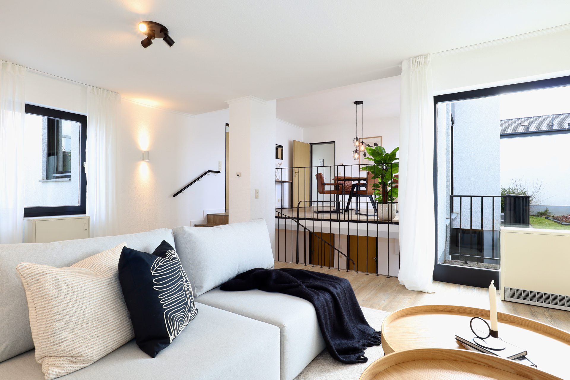 Ein modernes, minimalistisches Wohnzimmer mit hellgrauem Sofa, dunklen Zierkissen und freiem Blick in einen hellen Essbereich.