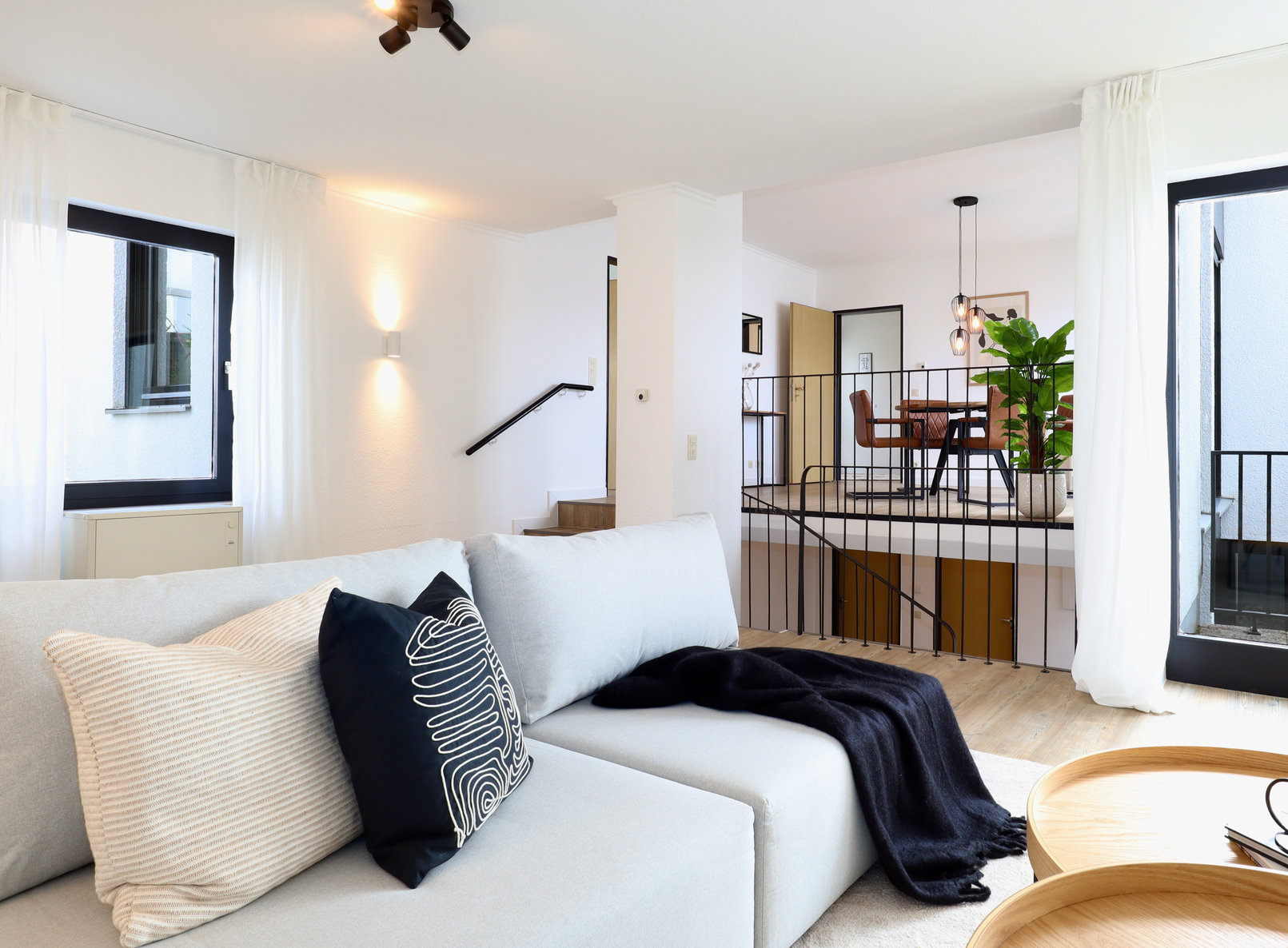 In einem modernen Wohnzimmer mit Blick auf einen tiefer gelegenen Essbereich steht ein helles Sofa mit einem Zierkissen.