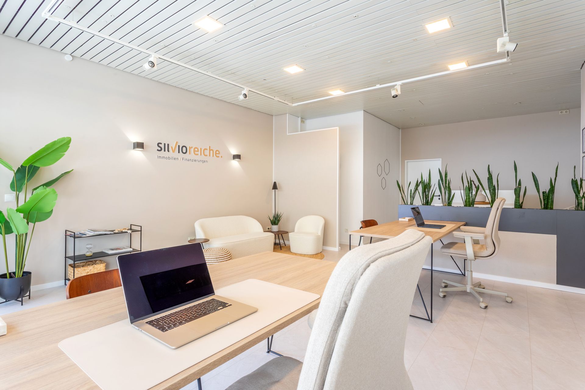 Ein modernes, minimalistisches Büro mit hellen Holzschreibtischen, einem weißen Sofa, Zimmerpflanzen.