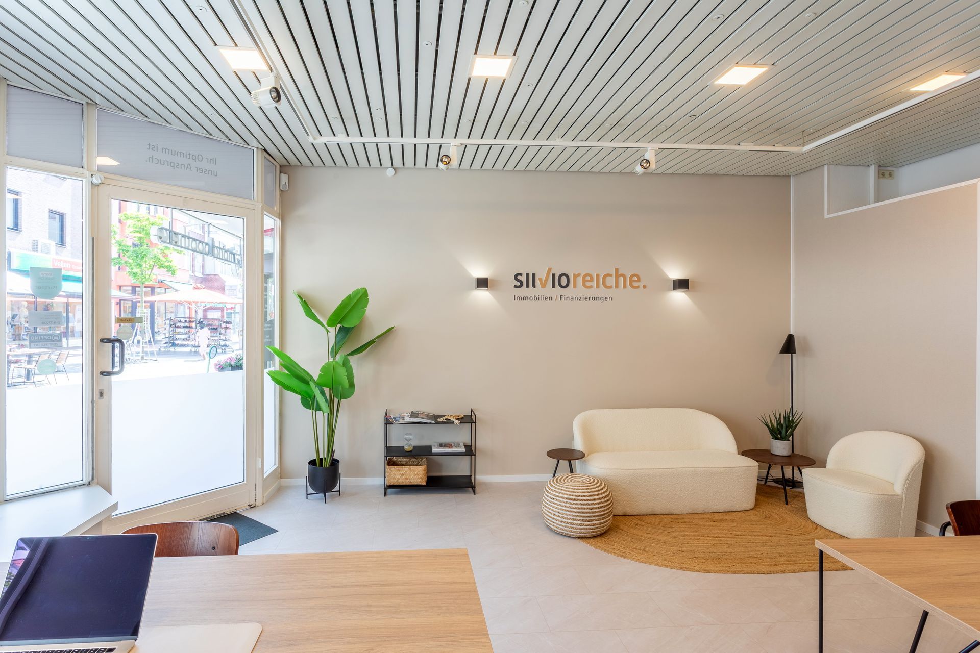 Ein moderner, minimalistischer Büroempfangsbereich mit Sofa, Sessel, großer Topfpflanze und an der Wand befestigtem Logo.