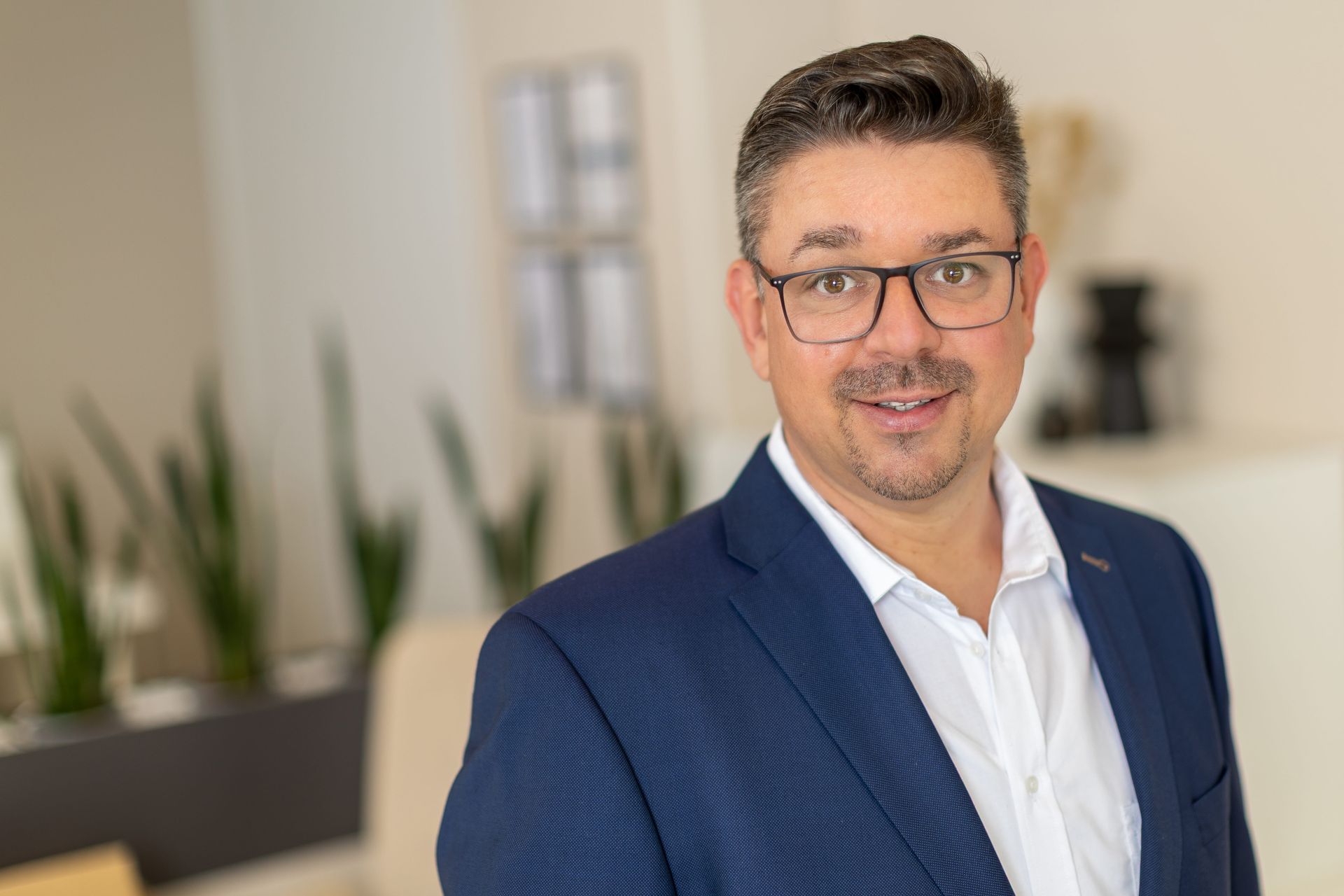 Ein professionelles Porträtfoto einer Person in einem blauen Blazer und mit Brille.