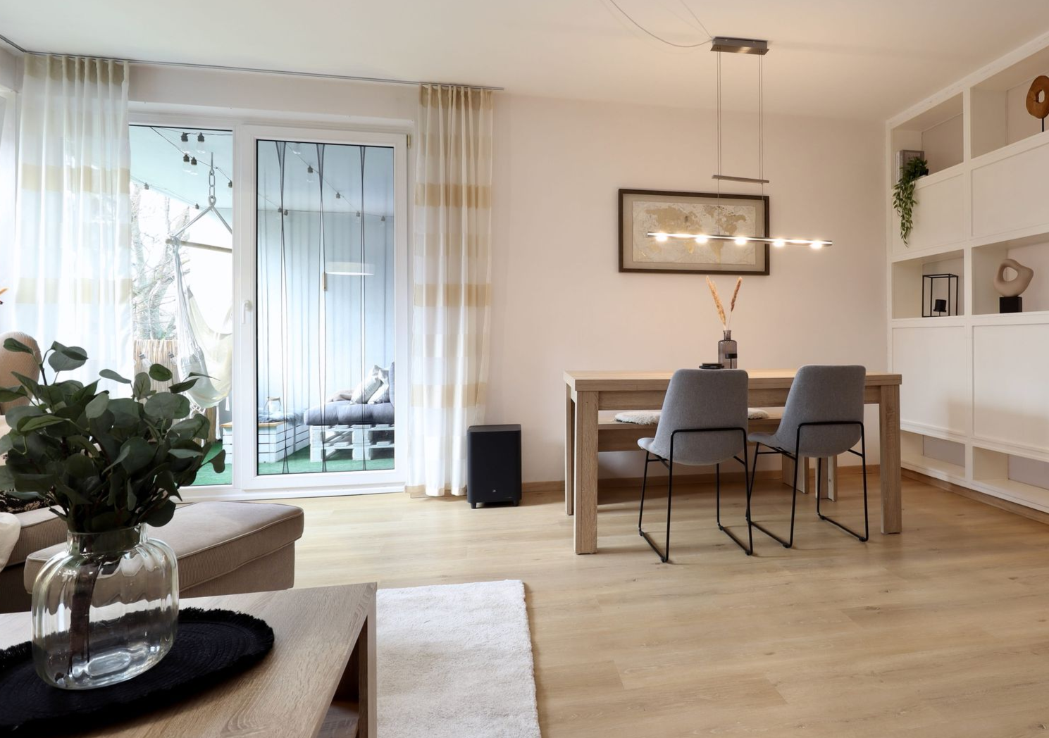 Ein heller, moderner Essbereich mit einem Holztisch, zwei grauen Stühlen, einer Pendelleuchte und großen Fenstern.