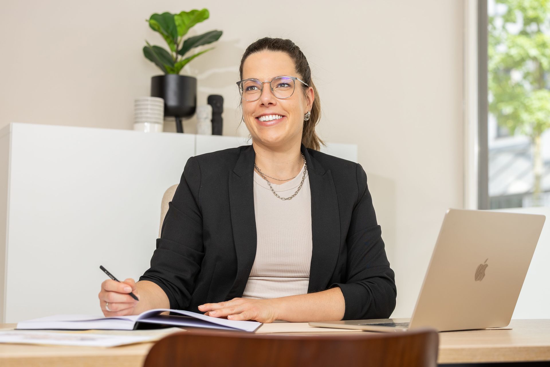 Eine Person in einem schwarzen Blazer lächelt, während sie an einem Schreibtisch im Büro.