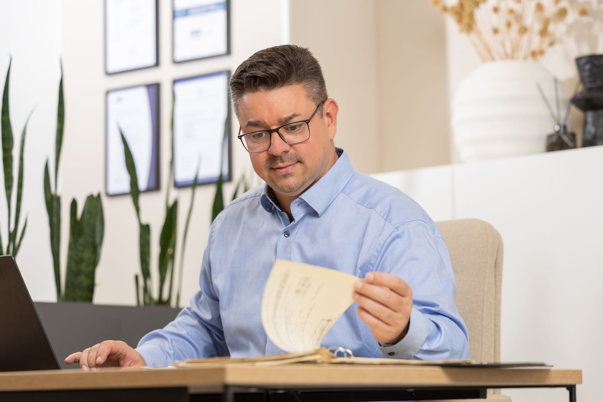 Eine Person in einem hellblauen Hemd mit Knöpfen und Brille sitzt an einem Schreibtisch und liest ein Dokument.