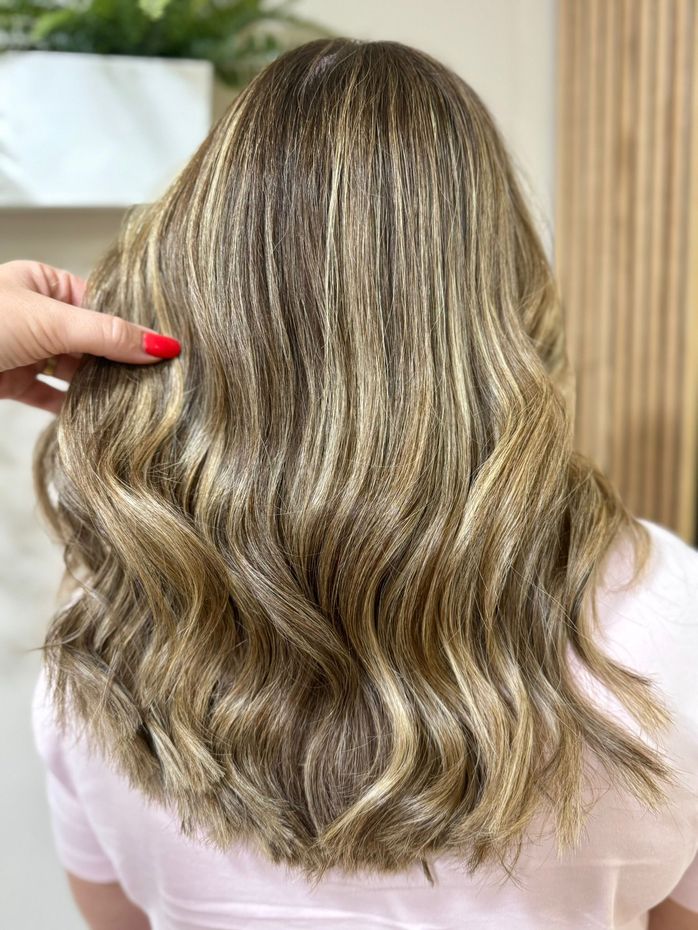 Mujer con cabello ondulado y mechas, en tonos castaños y rubios. Mano tocándose el cabello.