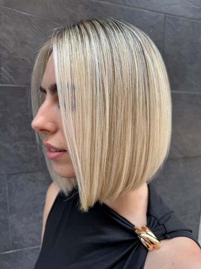 Mujer con corte de pelo bob rubio y recto, vestida con un top negro, posando frente a una pared gris.