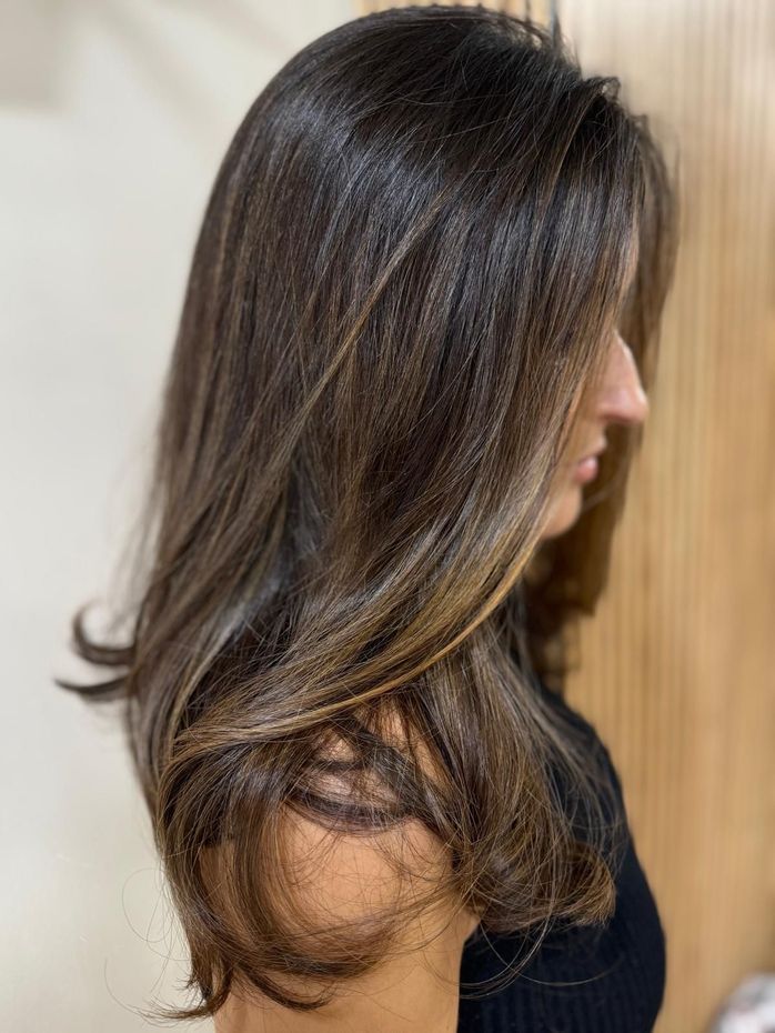 Mujer con cabello castaño largo, en capas y con reflejos. Perfil lateral, en interiores.