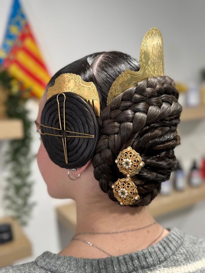 Mujer con peinado adornado, trenzas oscuras, tocado dorado y pendientes; bandera de Valencia al fondo.