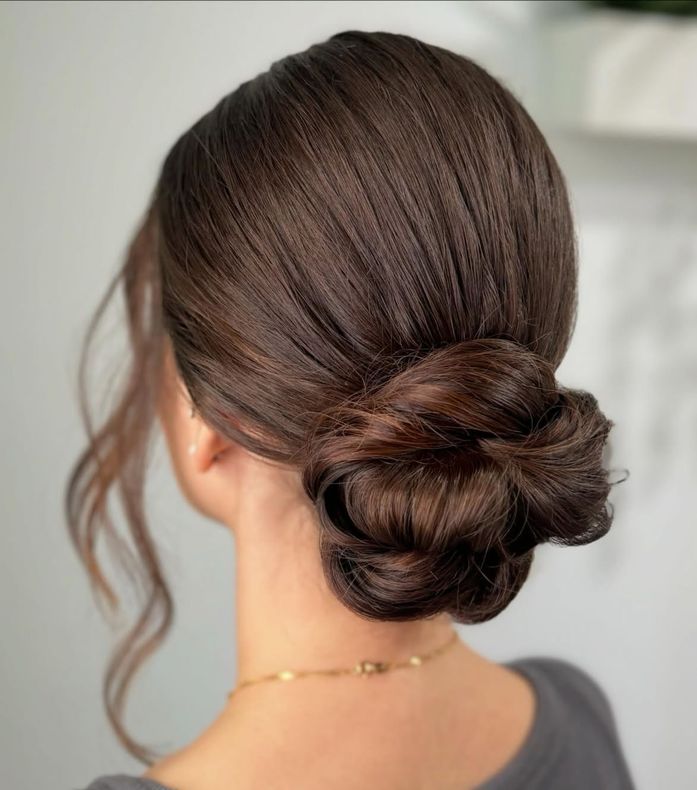 Mujer con cabello castaño oscuro recogido en un moño bajo con textura. Se ve un collar de oro.