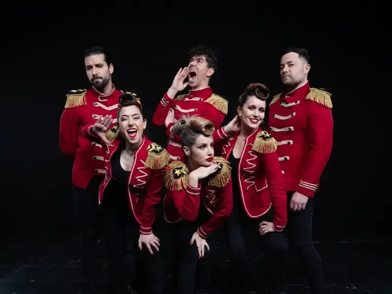 Seis artistas con chaquetas rojas de estilo militar posan sobre un fondo negro, sonriendo y gesticulando de forma juguetona.