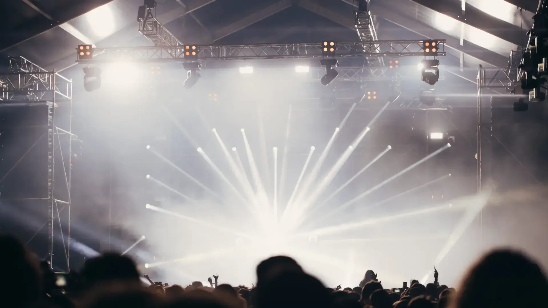 Escenario de conciertos con focos blancos brillantes y siluetas de público bajo un recinto techado.