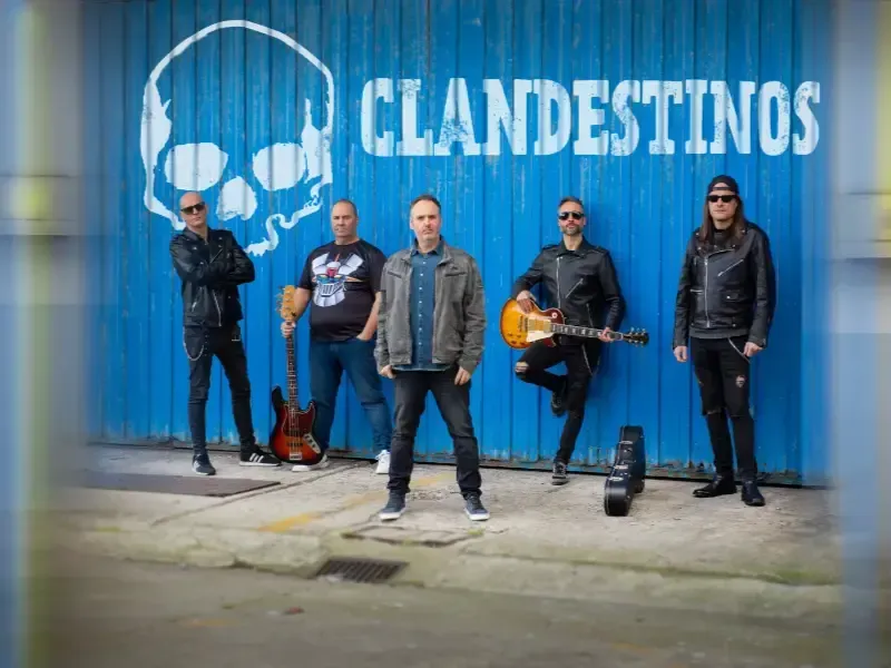 Cinco músicos se encuentran frente a una pared azul con el logo de una calavera y la palabra “CLANDESTINOS”.