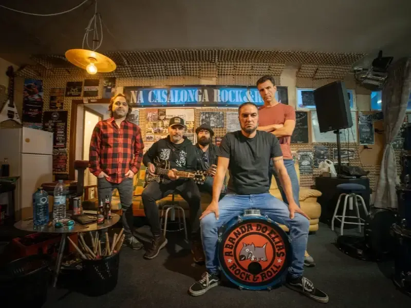 Cuatro personas en una sala de ensayo musical, con instrumentos y el logotipo de un parche de batería en primer plano.