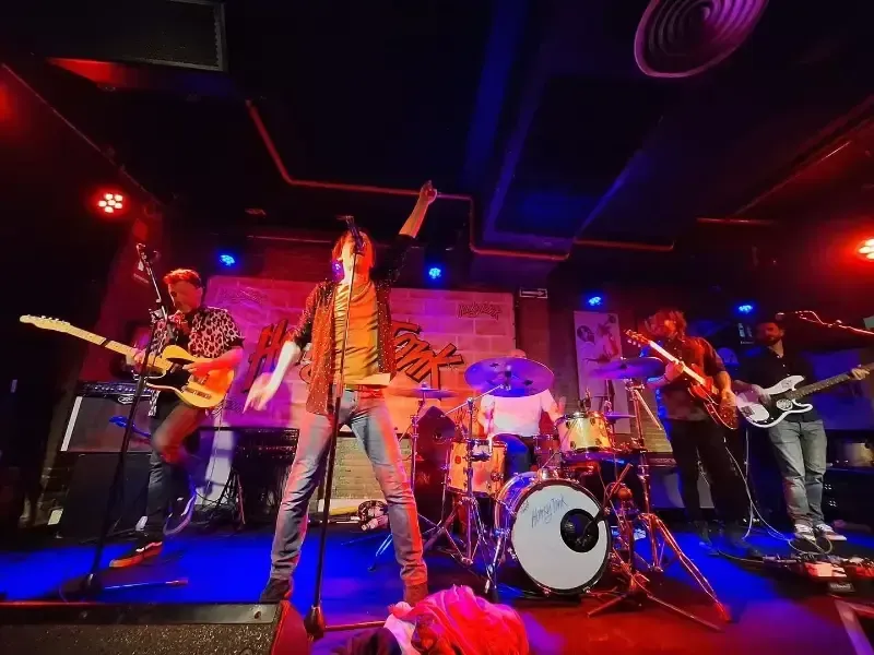 Banda actuando en directo en un escenario colorido con guitarras, batería e iluminación roja y azul.