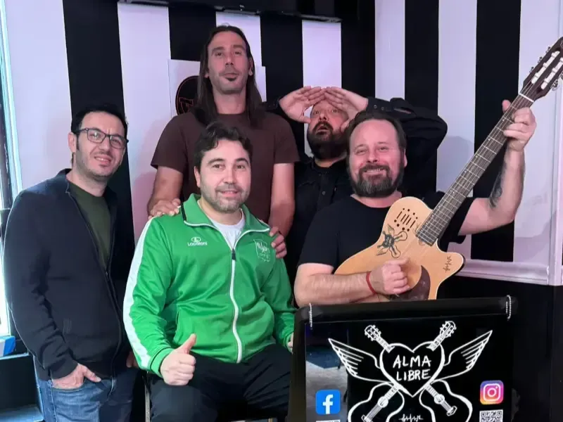 Cinco músicos posando en un estudio, uno de ellos con una guitarra, con un fondo de rayas blancas y negras.