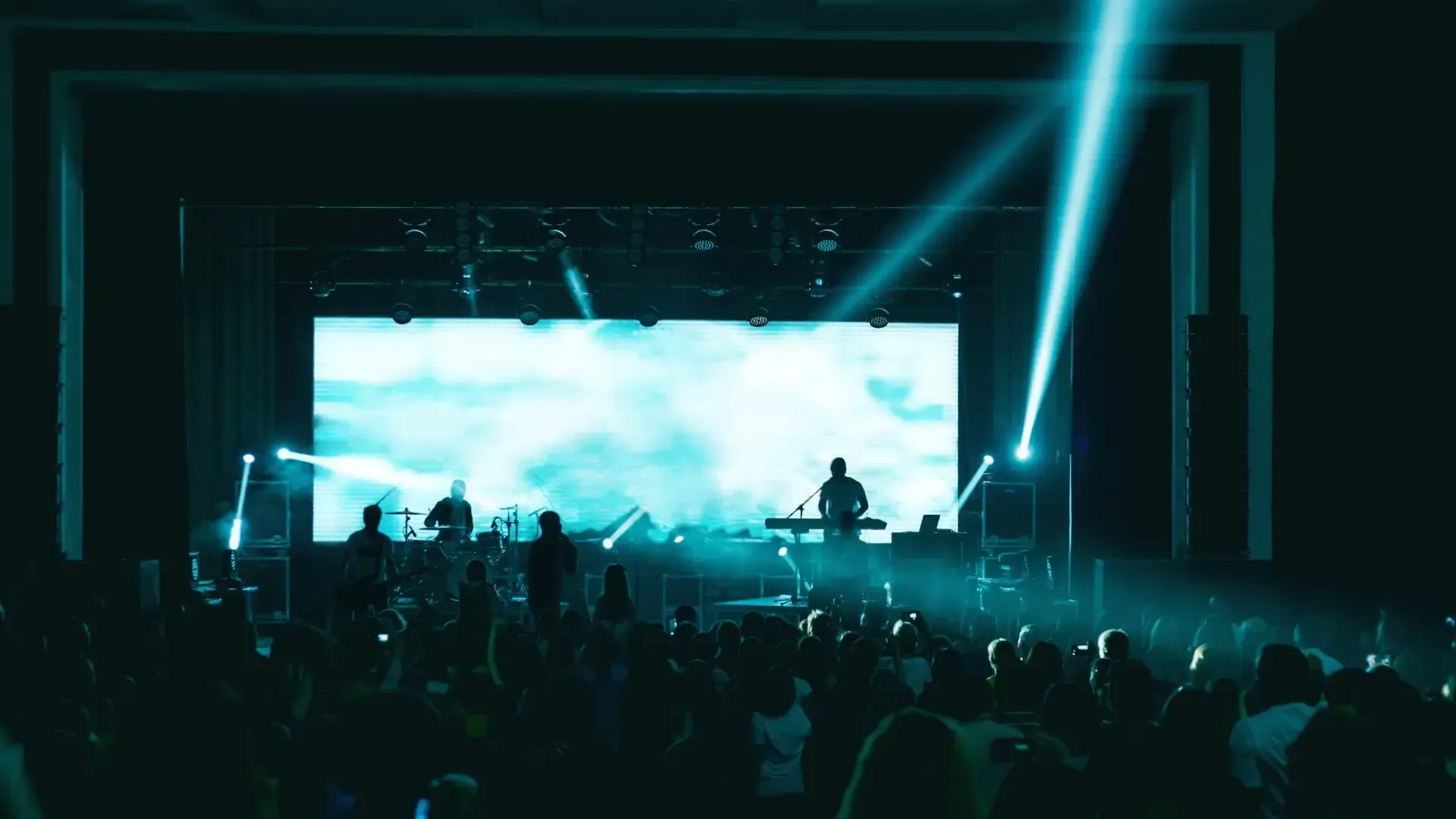 Escenario de concierto con luces azules brillantes, una gran pantalla y un artista en silueta frente a una multitud.