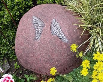 In einem Garten liegt ein großer Felsen mit eingeschnitzten Flügeln.