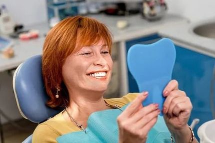 Una mujer está sentada en una silla dental mirándose los dientes en un espejo.