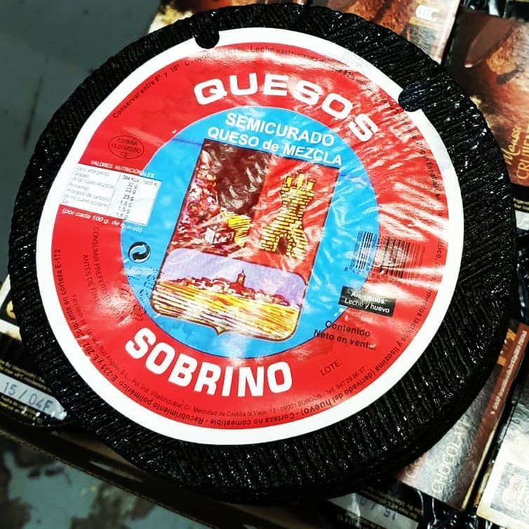 Una rueda de queso semicurado de leche mixta de la marca Sobrino, con corteza negra y etiqueta roja y azul.