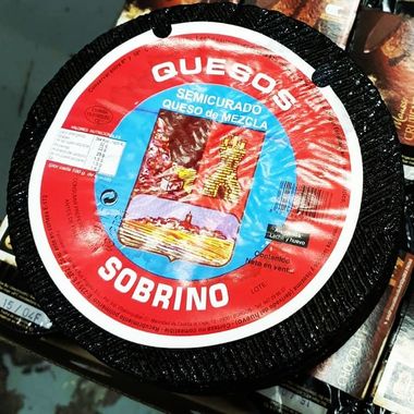 Una rueda de queso semicurado de leche mixta de la marca Sobrino, con corteza negra y etiqueta roja y azul.