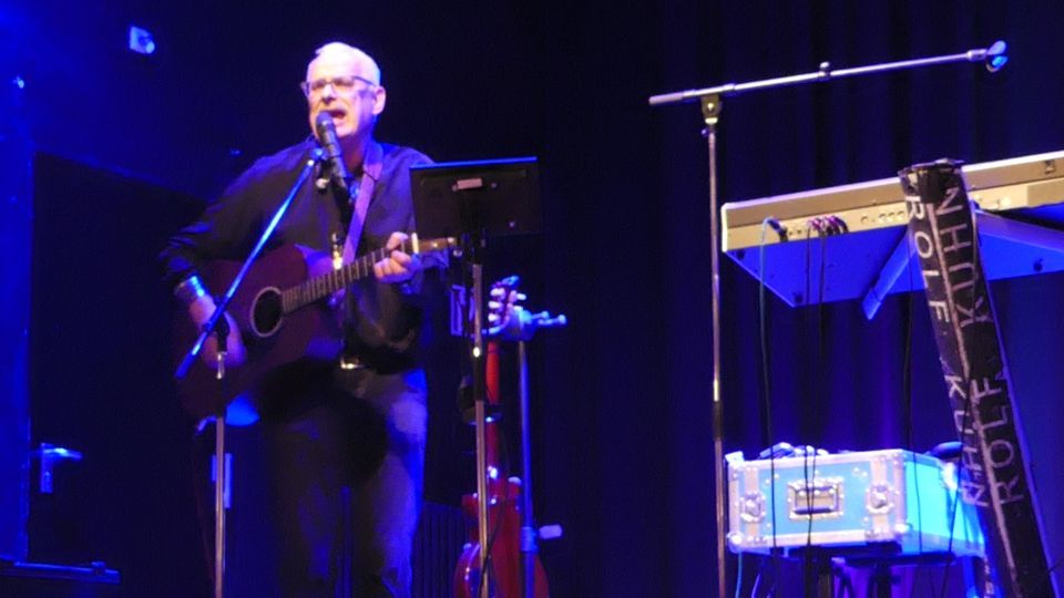 Auf einer Bühne spielt ein Mann Gitarre und singt in ein Mikrofon.