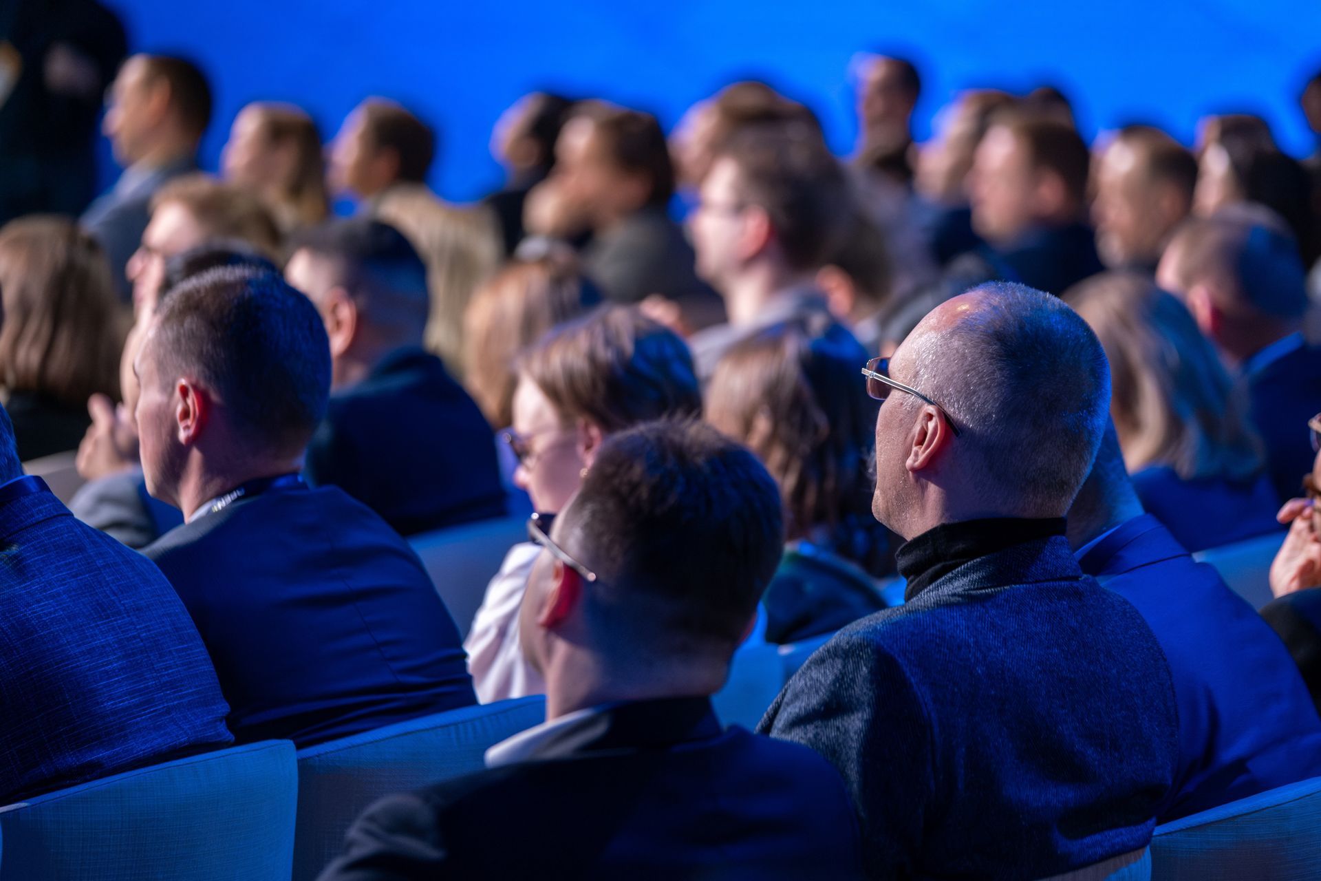 Zuschauer sitzen in einem abgedunkelten Auditorium, blicken nach vorne auf einen unsichtbaren Redner, vor einem blauen Hintergrund.