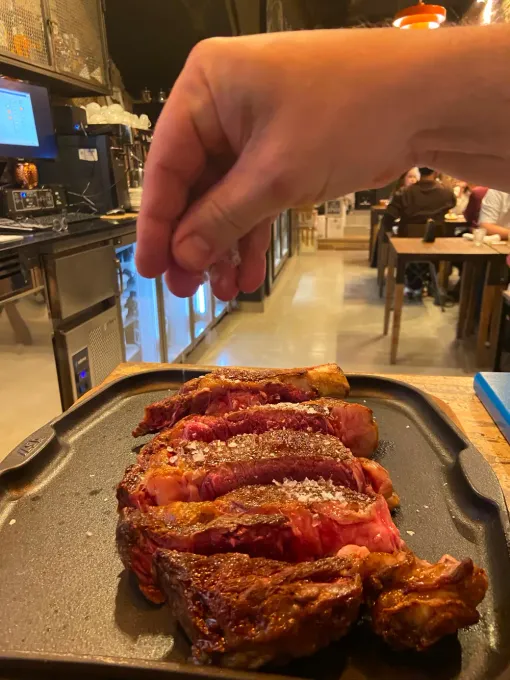 Una mano coloca un plato de carne asada sazonada sobre una mesa en la cocina de un restaurante.