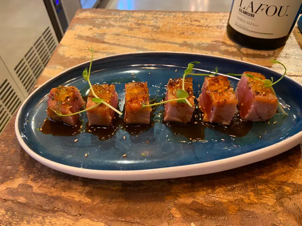 Plato azul con lonchas de atún sellado y microbrotes sobre una mesa de madera, con una botella de vino al fondo