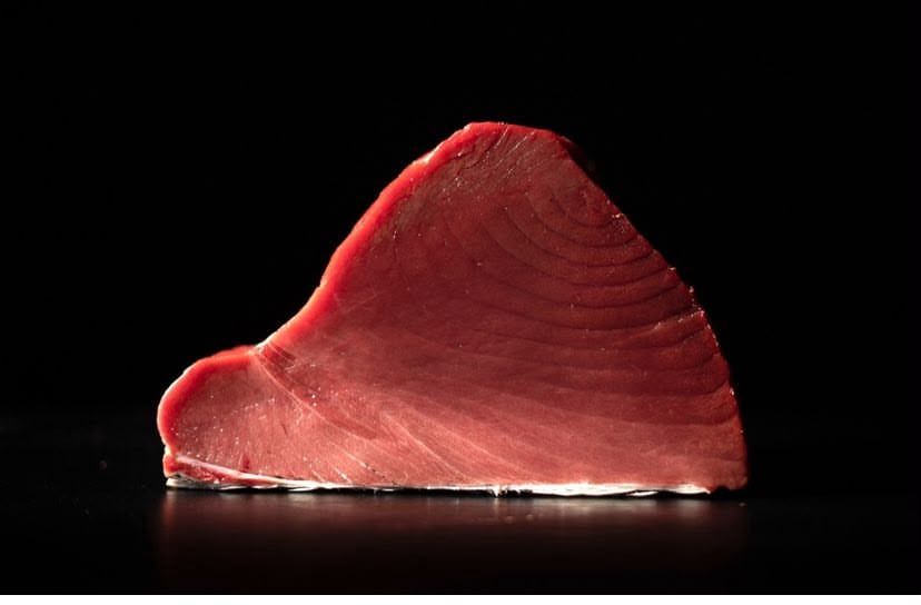 Un filete de pescado rosado y rojizo, cortado en lonchas y colocado en vertical sobre un fondo negro.